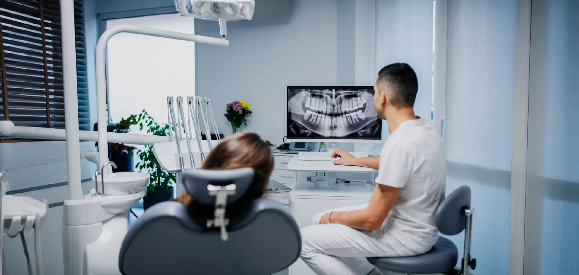 Man sieht einen Zahnarzt von hinten in einem Behandlungsraum mit einer Patientin. Sie schauen sich gemeinsam ein Röntgenbild an.